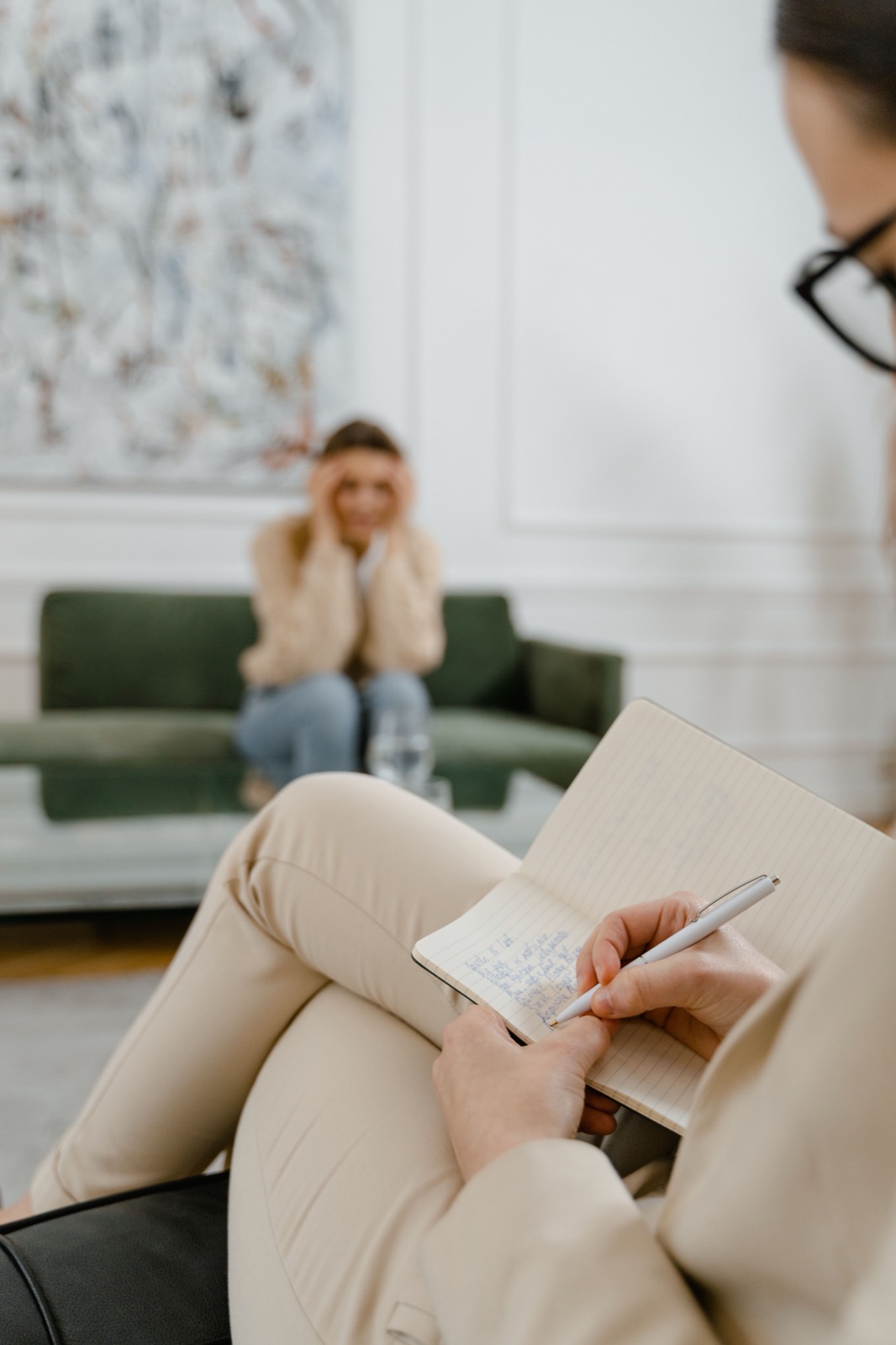 Een cliënt op de bank met de handen voor het gezicht; op de voorgrond maakt een behandelaar aantekeningen.